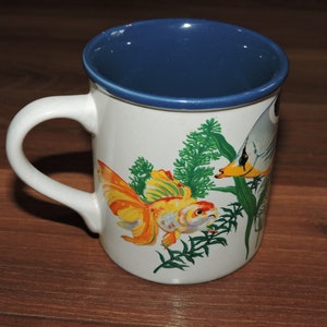 May include: White ceramic mug with a blue rim and a colourful illustration of goldfish and coral. The mug has a handle.