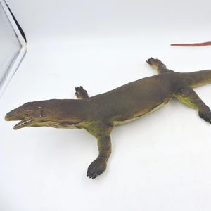 May include: A detailed, brown and green lizard figurine with a long, red tail. The reptile has visible claws and an open mouth, showcasing its teeth. The toy is set against a white background, highlighting its features.