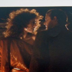 May include: A woman in a long, orange dress stands near a man in a dark suit. They are looking at each other, and the woman is holding a bouquet of flowers.