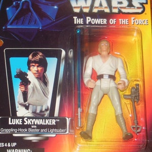Op de afbeelding: Een plastic actieve figuur van Luke Skywalker uit Star Wars: The Power of the Force. De figuur draagt een wit pak en houdt een lichtzwaard vast. De figuur is verpakt in een doorzichtige plastic blisterverpakking met een gele en rode achtergrond.