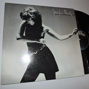 Op de afbeelding: Een zwart-witfoto van een vrouw in een zwarte jurk en een riem. De foto is op een vinyl platenhoes met de tekst "Jennie Rush" geschreven in zwarte inkt.