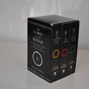 May include: A black boxed set of The Lord of the Rings books by J.R.R. Tolkien. The box features a silver ring with the inscription "The Fellowship of the Ring, The Two Towers, The Return of the King". The individual books have colored rings on the spines: yellow, red, and green.