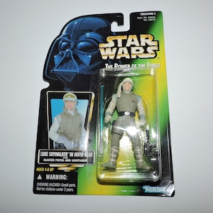 Può includere: Una action figure di Star Wars di Luke Skywalker in equipaggiamento Hoth, che tiene una pistola blaster e una spada laser. La figura è confezionata in una scatola di cartone verde e gialla con il logo di Star Wars e le parole "The Power of the Force".