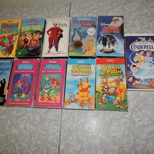 Pu&ograve; includere: Una collezione di videocassette VHS Disney con classici animati come "Cenerentola", "La Sirenetta" e "Winnie the Pooh". Altri titoli includono "Cip & Ciop: Double Trouble" e "The Santa Clause". Le cassette sono disposte su una superficie bianca.
