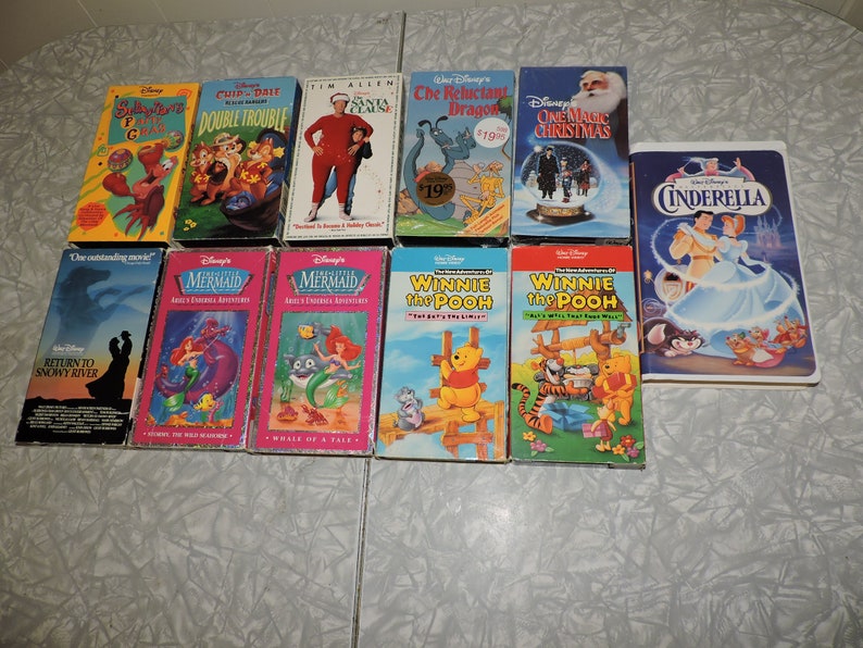 Pu&ograve; includere: Una collezione di videocassette VHS Disney con classici animati come "Cenerentola", "La Sirenetta" e "Winnie the Pooh". Altri titoli includono "Cip & Ciop: Double Trouble" e "The Santa Clause". Le cassette sono disposte su una superficie bianca.