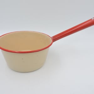 Puede incluir: Cacerola de esmalte color crema con borde y asa rojos. El asa se extiende desde el lateral de la cacerola y el borde es de color rojo brillante. La cacerola está vacía y se encuentra sobre una superficie blanca.
