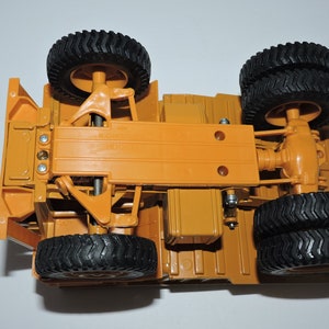May include: Underside view of a toy construction vehicle with four black wheels and a yellow chassis. The vehicle has a detailed suspension system and a visible rear axle.