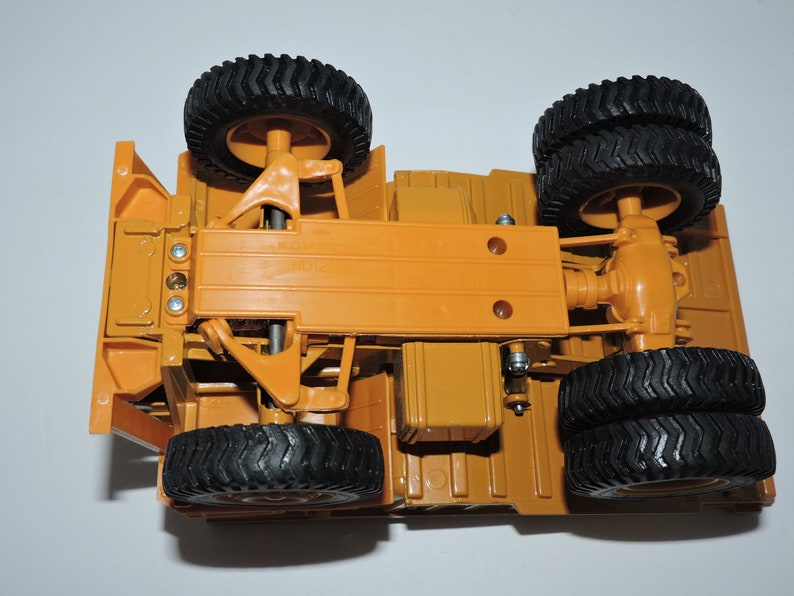 May include: Underside view of a toy construction vehicle with four black wheels and a yellow chassis. The vehicle has a detailed suspension system and a visible rear axle.