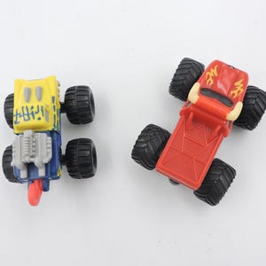 May include: Two toy monster trucks. One is yellow and blue with black wheels, the other is red with black wheels. The trucks have large, textured tires and detailed designs on the bodies.