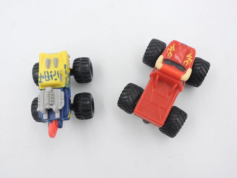 May include: Two toy monster trucks. One is yellow and blue with black wheels, the other is red with black wheels. The trucks have large, textured tires and detailed designs on the bodies.
