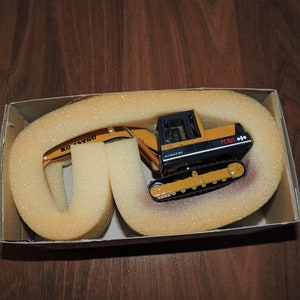 May include: A yellow and black toy excavator with the text "KOMATSU" and "PC100" on the side. The excavator is in a cardboard box with foam padding.