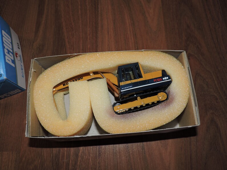 May include: A yellow and black toy excavator with the text "KOMATSU" and "PC100" on the side. The excavator is in a cardboard box with foam padding.