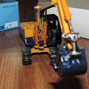 May include: A yellow toy excavator with a black cab and a blue bucket. The excavator is on a wooden surface and the box it came in is visible in the background. The box says "PC100 D SERIES" and "MADE IN JAPAN".