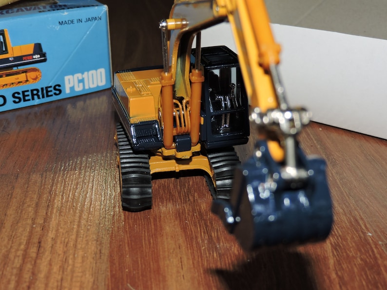 May include: A yellow toy excavator with a black cab and a blue bucket. The excavator is on a wooden surface and the box it came in is visible in the background. The box says "PC100 D SERIES" and "MADE IN JAPAN".