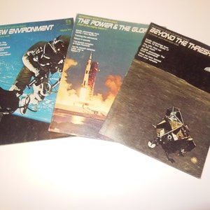 Op de afbeelding: Drie vintage paperbacks over ruimteverkenning. De omslagen tonen een ruimtewandeling, een raketlancering en een maanlanding. De titels zijn "Een nieuwe omgeving", "De kracht en de bol" en " voorbij de drempel".