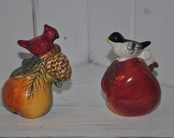 May include: A pair of ceramic salt and pepper shakers shaped like pears. The pear on the left has a red cardinal perched on a pine cone, while the pear on the right has a black and white bird perched on a pine branch.