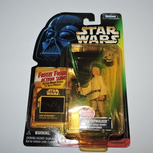 Op de afbeelding: Een Star Wars-actiefiguur van Luke Skywalker in zijn Jedi Knight-outfit, met een lightsaber in de hand. De figuur is verpakt in een plastic blisterverpakking met een gouden achtergrond en een freeze frame-action slide. De verpakking bevat ook het Star Wars-logo en de woorden "The Power of the Force".