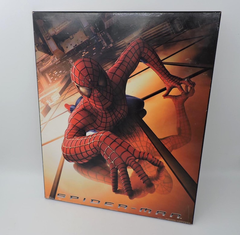 Op de afbeelding: Een ingelijste afbeelding met Spider-Man in zijn rood-blauwe pak, die zich aan een gebouw vastklampt. Het kunstwerk heeft een warme, oranje en gele achtergrond met de woorden "SPIDER-MAN" onderaan.
