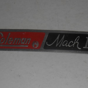 Peut inclure: Un badge en métal argenté avec les mots "Coleman" en rouge et "Mach II" en noir.
