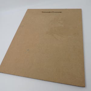 Op de afbeelding: Een rechthoekige, bruine kartonnen achterplaat met een klein gaatje aan de bovenkant om op te hangen. De plaat heeft een licht getextureerd oppervlak en is in de afbeelding schuin geplaatst. De achtergrond is wit.