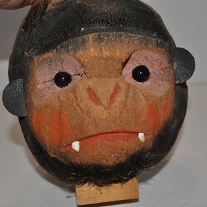 May include: A carved coconut monkey head with black hair, painted eyes, and a red mouth. The monkey is mounted on a wooden base.