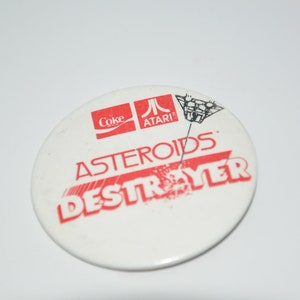 Puede incluir: Un botón redondo blanco con texto rojo que dice "Asteroids Destroyer" y una imagen en blanco y negro de una nave espacial. El botón también tiene los logotipos de Coca-Cola y Atari.