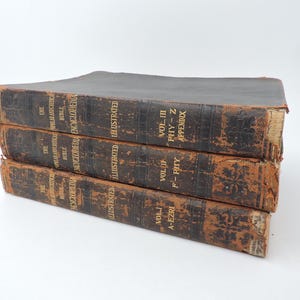 May include: Three antique, leather-bound books stacked on top of each other. The books are dark brown with gold lettering and feature the title "The Philadelphia Bible Encyclopedia Illustrated." The edges of the books show signs of wear.