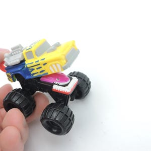 May include: A colorful toy monster truck with large black tires. The truck's cab is yellow with blue and gray accents, and a shark-themed front end. The toy is held in a hand against a white background.