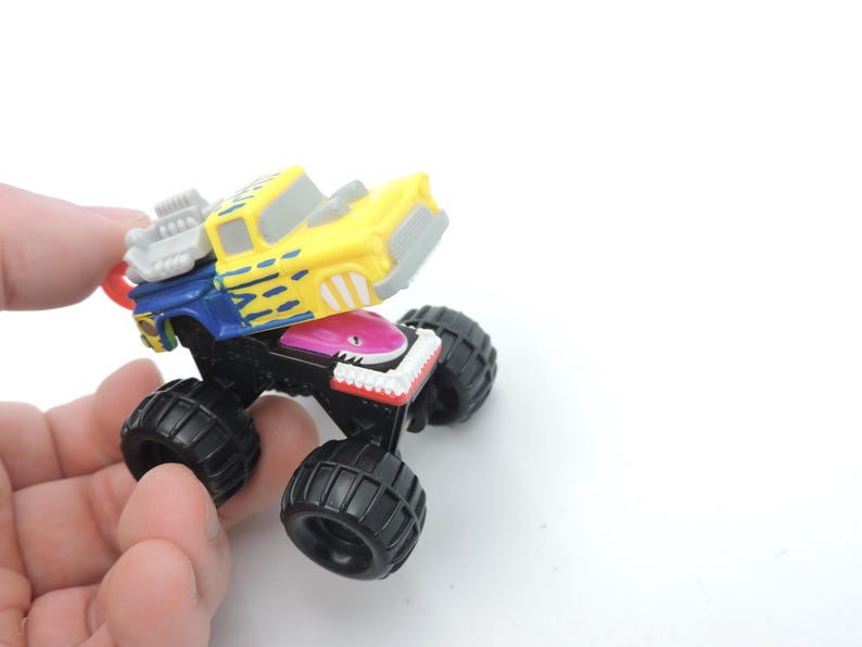 May include: A colorful toy monster truck with large black tires. The truck's cab is yellow with blue and gray accents, and a shark-themed front end. The toy is held in a hand against a white background.