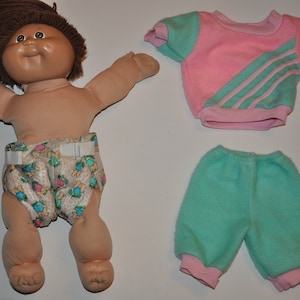 Puede incluir: Una muñeca vintage con cabello castaño, tez bronceada y un pañal con estampado floral. La muñeca está acompañada de una sudadera rosa y verde azulado y pantalones verde azulado con puños rosas. Un juego de juguetes nostálgico.