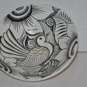 May include: A white ceramic bowl with a black and white design of a bird with a large, feathery tail. The bowl has a scalloped edge and the words "Columel" and "Mon" are written on the side.