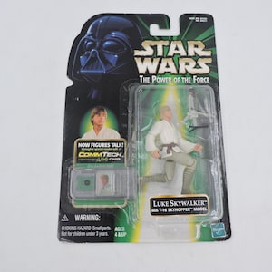 Op de afbeelding: Een Star Wars "The Power of the Force" actiefiguur van Luke Skywalker met een T-16 Skyhopper model, in de originele verpakking. De verpakking toont Darth Vader, het Star Wars logo en de tekst "Now Figures Talk!" en "CommTech Chip."