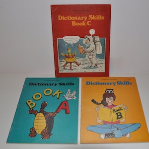 Puede incluir: Tres libros de bolsillo coloridos titulados "Dictionary Skills" con diferentes ilustraciones en cada portada. El libro A presenta una tortuga sosteniendo una letra A, el libro B presenta a una persona leyendo un libro con un mapache y el libro C presenta a un astronauta y un extraterrestre.