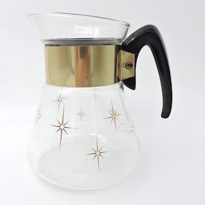 Peut inclure: Une carafe en verre transparent avec une bande dorée et une poignée noire. La carafe est décorée de motifs d'éclats d'étoiles dorées. La carafe est probablement utilisée pour servir des boissons.
