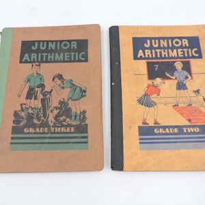 Peut inclure: Deux manuels scolaires anciens intitulés Junior Arithmetic. Le livre de gauche est marron avec une tranche verte et marqué Grade Three, illustré d'un garçon et d'une fille. Le livre de droite est beige avec une tranche noire et marqué Grade Two.