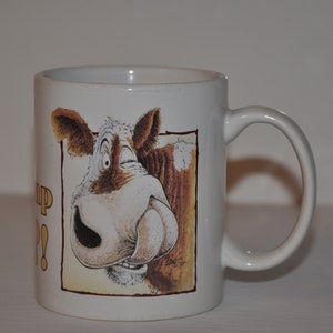 Op de afbeelding: Witte keramische mok met een cartoon-koeiengezichts-illustratie. De koe kijkt de kijker aan met een speelse uitdrukking. De tekst "Moo Up?" staat gedrukt op de zijkant van de mok.