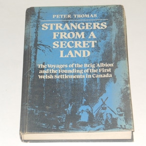 May include: A blue book cover with the title "Strangers From A Secret Land" by Peter Thomas. The subtitle is "The Voyages of the Brig Albion and the Founding of the First Welsh Settlements in Canada".