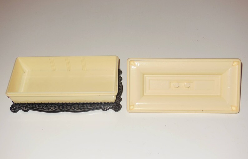 May include: A rectangular cream-colored box with a black ornate base and a matching lid. The lid has two small round buttons.