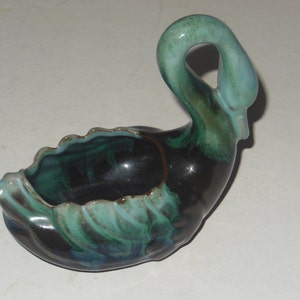 Puede incluir: Un macetero de cerámica en forma de cisne, esmaltado en negro y verde. El cisne tiene un cuello largo y un pico ancho y abierto. El macetero tiene forma de cisne con un cuerpo hueco.
