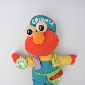 May include: A plush toy of a red character with large, round eyes, an orange nose, and a blue cap that says "CRINKLE". The character wears a yellow shirt, blue overalls, and a green fanny pack with a red zipper. A green circle on the left arm says "SNAP".