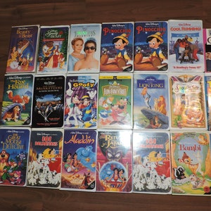 Pu&ograve; includere: Una collezione di VHS Disney, con titoli come "La Bella e la Bestia" e "Il Re Leone". Le cassette sono disposte su una superficie di legno, con illustrazioni colorate e titoli di film.