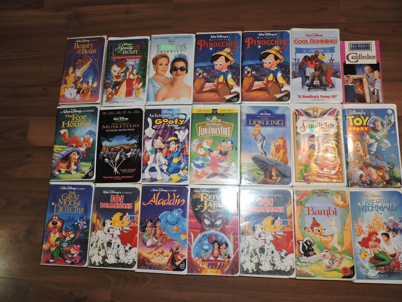 Pu&ograve; includere: Una collezione di VHS Disney, con titoli come "La Bella e la Bestia" e "Il Re Leone". Le cassette sono disposte su una superficie di legno, con illustrazioni colorate e titoli di film.