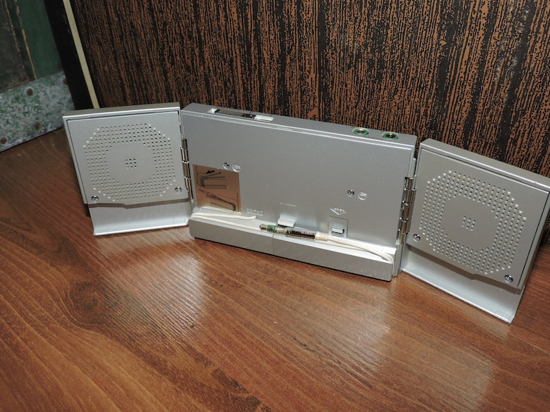 May include: A silver-colored portable speaker system with three sections. The center section has a screen and ports. The two side sections have speakers with a grill pattern.