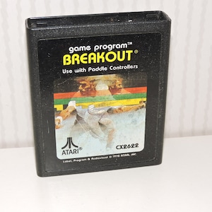 Könnte beinhalten: Eine schwarze Atari-Spielkassette mit einem gelben Etikett, auf dem "game program™ BREAKOUT® Use with Paddle Controllers" steht. Das Etikett zeigt auch eine Grafik von zwei Personen, die das Spiel spielen. Die Kartuschennummer ist CX2622.