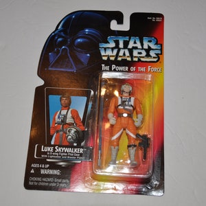 Op de afbeelding: Een Star Wars-actiefiguur van Luke Skywalker in X-wing-pilotenkleding met een lightsaber en een blasterpistool. De verpakking heeft het Star Wars-logo en de tekst "The Power of the Force".