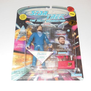 Op de afbeelding: Een Star Trek: The Next Generation actionfiguur van Commander Riker in zijn blauwe uniform. De figuur houdt een phaser vast en staat op een blauwe en witte basis. De verpakking bevat een Starfleet-uitrusting accessoire en een kaart met een foto van Riker en de tekst "Star Trek: The Next Generation" en "Ruimtecommando".