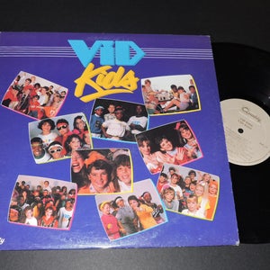 May include: A vinyl record album cover featuring a group of children on the front. The album is titled "Vid Kids" and features a blue and yellow colour scheme. The album cover is surrounded by a black vinyl record.