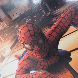 Op de afbeelding: Close-up van Spider-Man in zijn rood-zwarte webkostuum, met een stadsgezicht op de achtergrond. De superheld bevindt zich in een dynamische pose, met zijn hoofd en arm op de voorgrond.