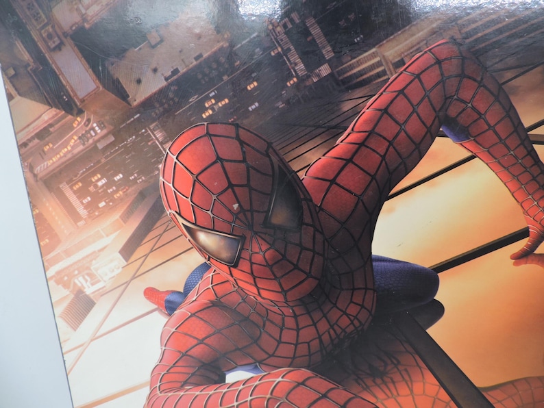 Op de afbeelding: Close-up van Spider-Man in zijn rood-zwarte webkostuum, met een stadsgezicht op de achtergrond. De superheld bevindt zich in een dynamische pose, met zijn hoofd en arm op de voorgrond.