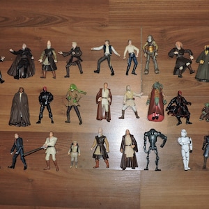 Op de afbeelding: Een verzameling Star Wars-actiefiguren, met verschillende personages in verschillende poses. De plastic figuren zijn in verschillende kleuren, waaronder zwart, wit, bruin en goud. De figuren zijn gerangschikt op een houten oppervlak.